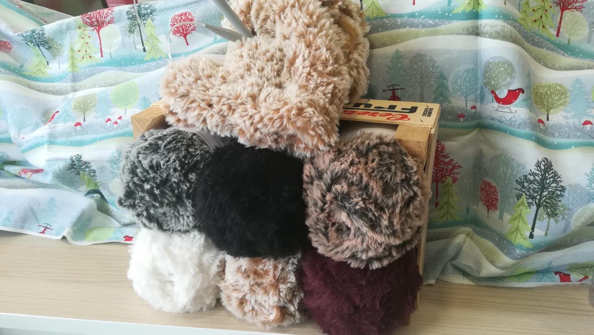 Novelos de lã felpuda multicolor em superfície clara com tecido estampado de árvores e casas ao fundo