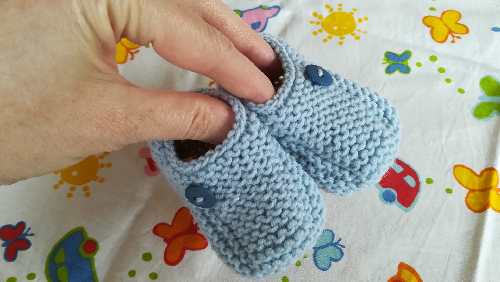 Sapatinhos para bebé azul claro em malha com botões azuis sobre tecido branco com desenhos coloridos
