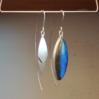 Brincos pendurados com metal prateado e padrão azul iridescente em fundo neutro.