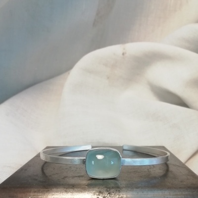 Pulseira prateada com pedra azul esverdeada sobre superfície metálica e pano branco