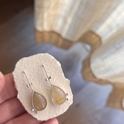 Brincos com pedras amarelas em forma de gota, base prateada sobre base branca irregular.