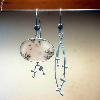 Par de brincos prateados com pedra branca e manchas pretas e detalhes azuis