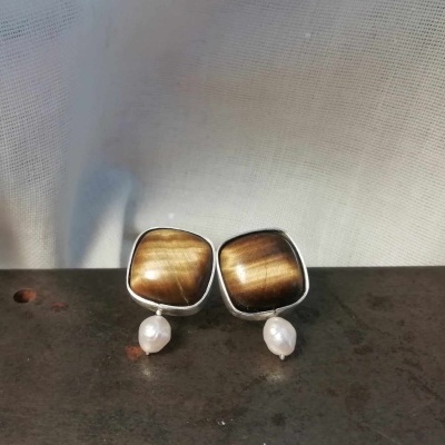 Brincos com pedras castanho dourado e pérolas pendentes, sobre superfície metálica escura.