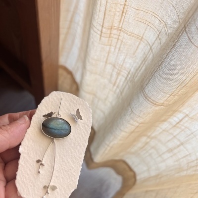 Brinco pendente com pedra oval azul em metal prateado e cartão texturizado ao fundo cortina branca.