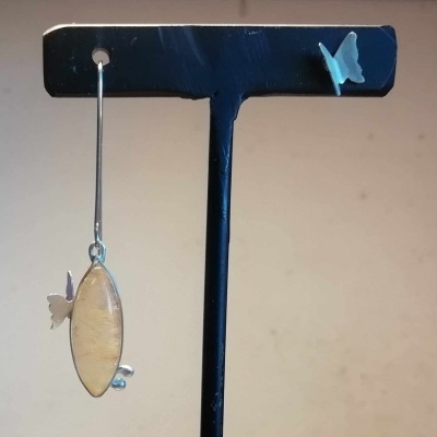 Brinco pendente oval prateado com borboleta e pérolas, em suporte preto com borboleta azul.