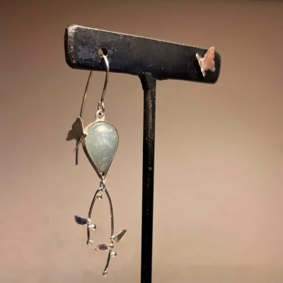 Brinco prateado com pedra verde e detalhes borboleta pendurado em suporte preto.