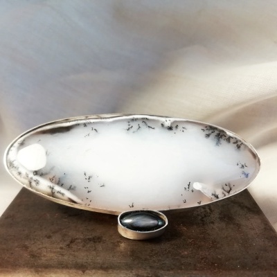 Broche oval branco com inclusões pretas e suporte metálico prateado