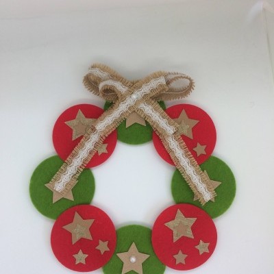 Coroa decorativa em feltro com círculos vermelhos e verdes, estrelas douradas e laço de juta com renda branca