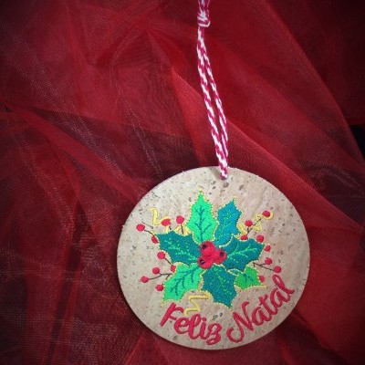 Ornamento de Natal redondo em cortiça com folhas verdes, bagas vermelhas e texto Feliz Natal