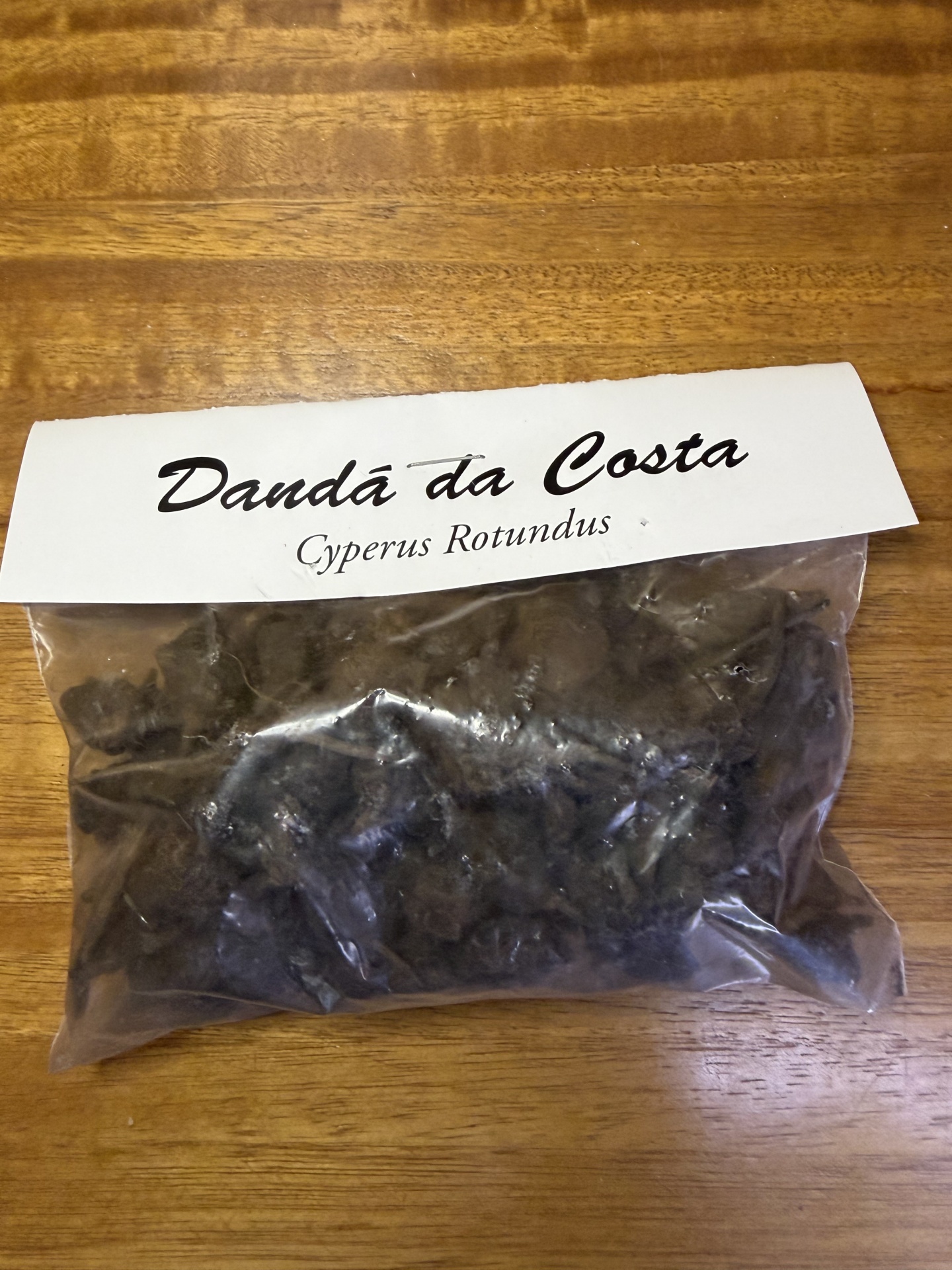 Embalagem plástica com pó escuro e etiqueta Dandá da Costa Cyperus Rotundus sobre mesa de madeira