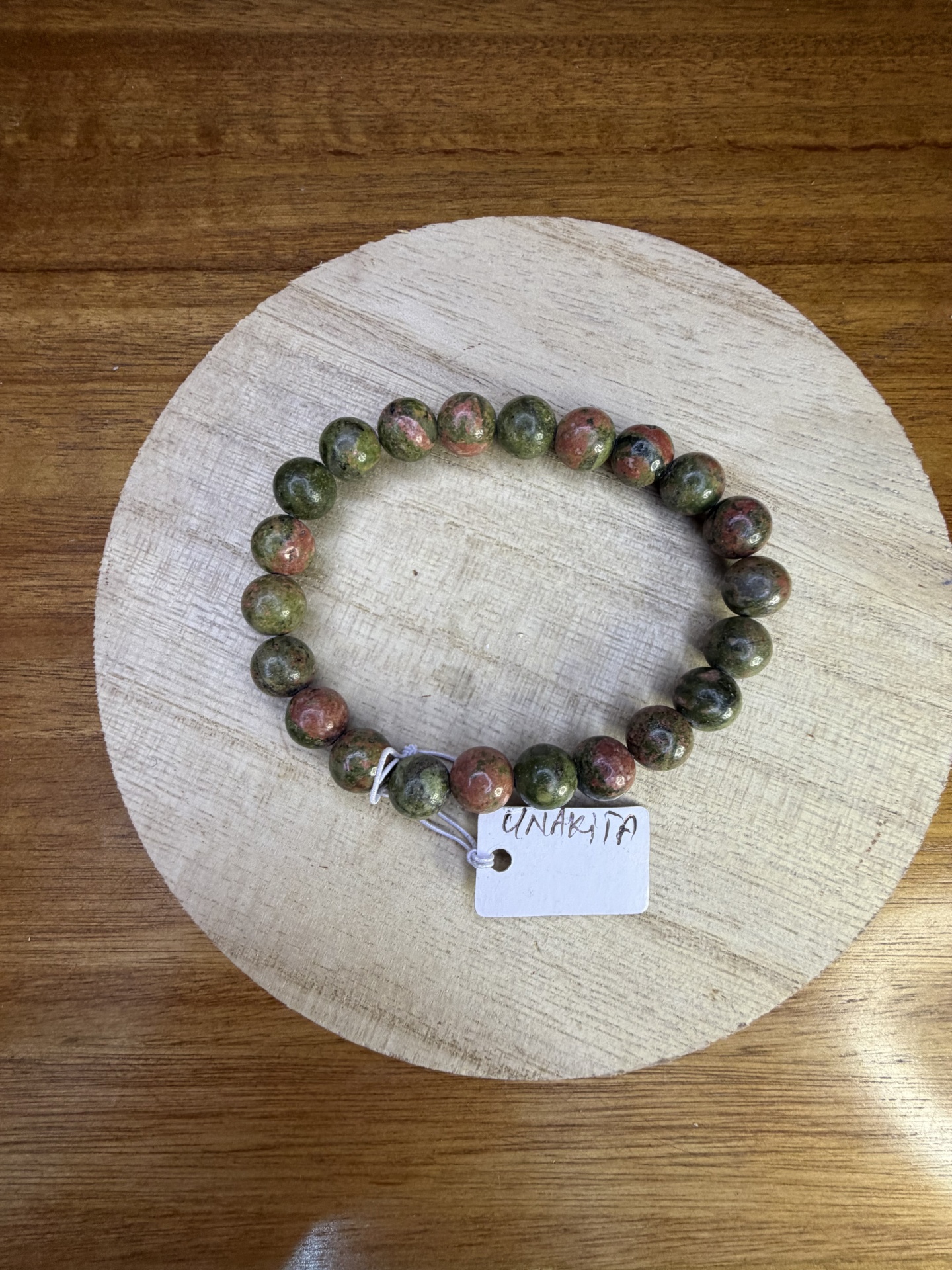 Pulseira de pedra verde e rosa com etiqueta UNAKITA numa superfície de madeira.