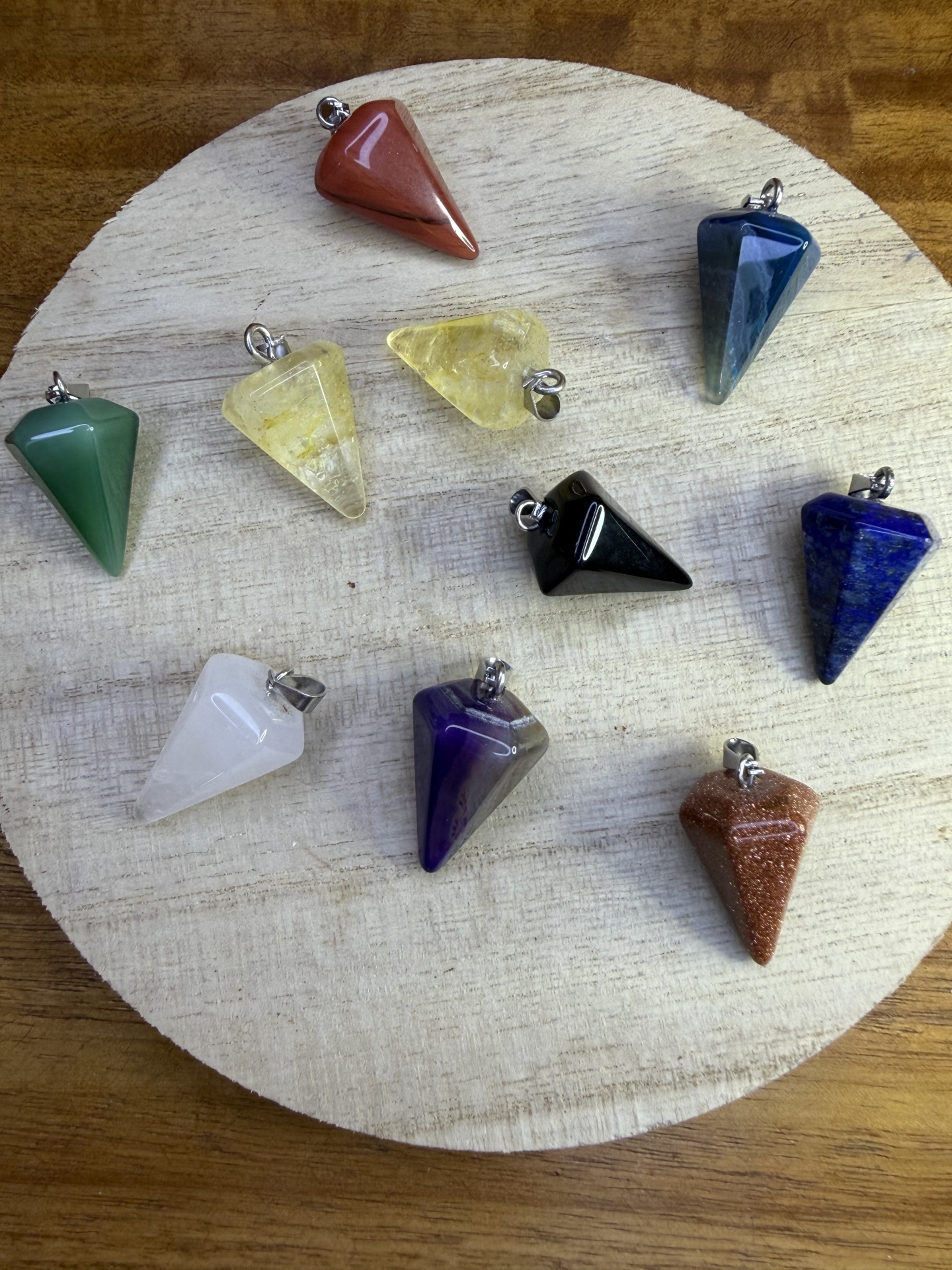 Dez pendentes de pedra polida coloridos com argolas metálicas, dispostos sobre superfície redonda clara