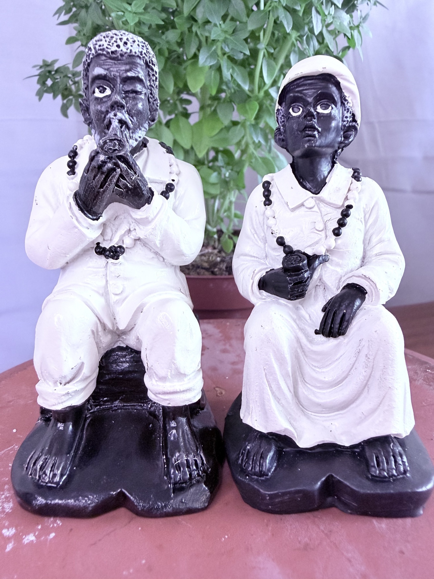 Esculturas em preto e branco de duas figuras humanas sentadas com colares, em frente a planta verde.