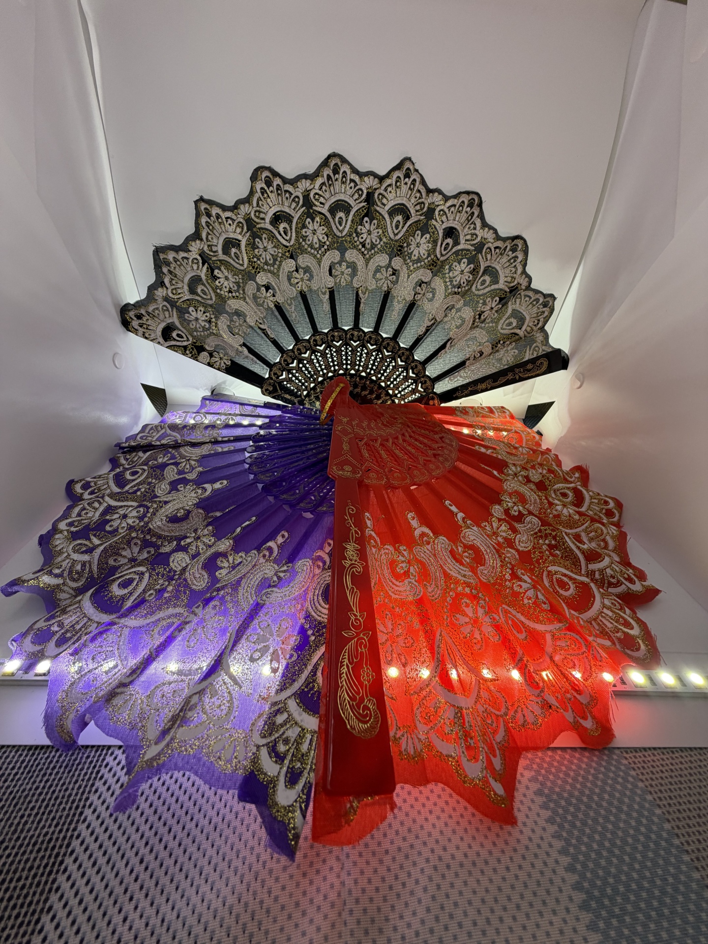 Três leques decorativos coloridos com padrões dourados e pretos numa parede branca