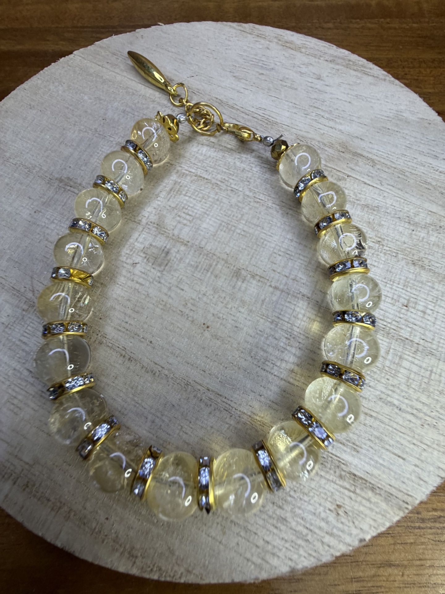 Pulseira de contas transparentes amareladas com aros dourados e pedras brilhantes sobre base de madeira clara
