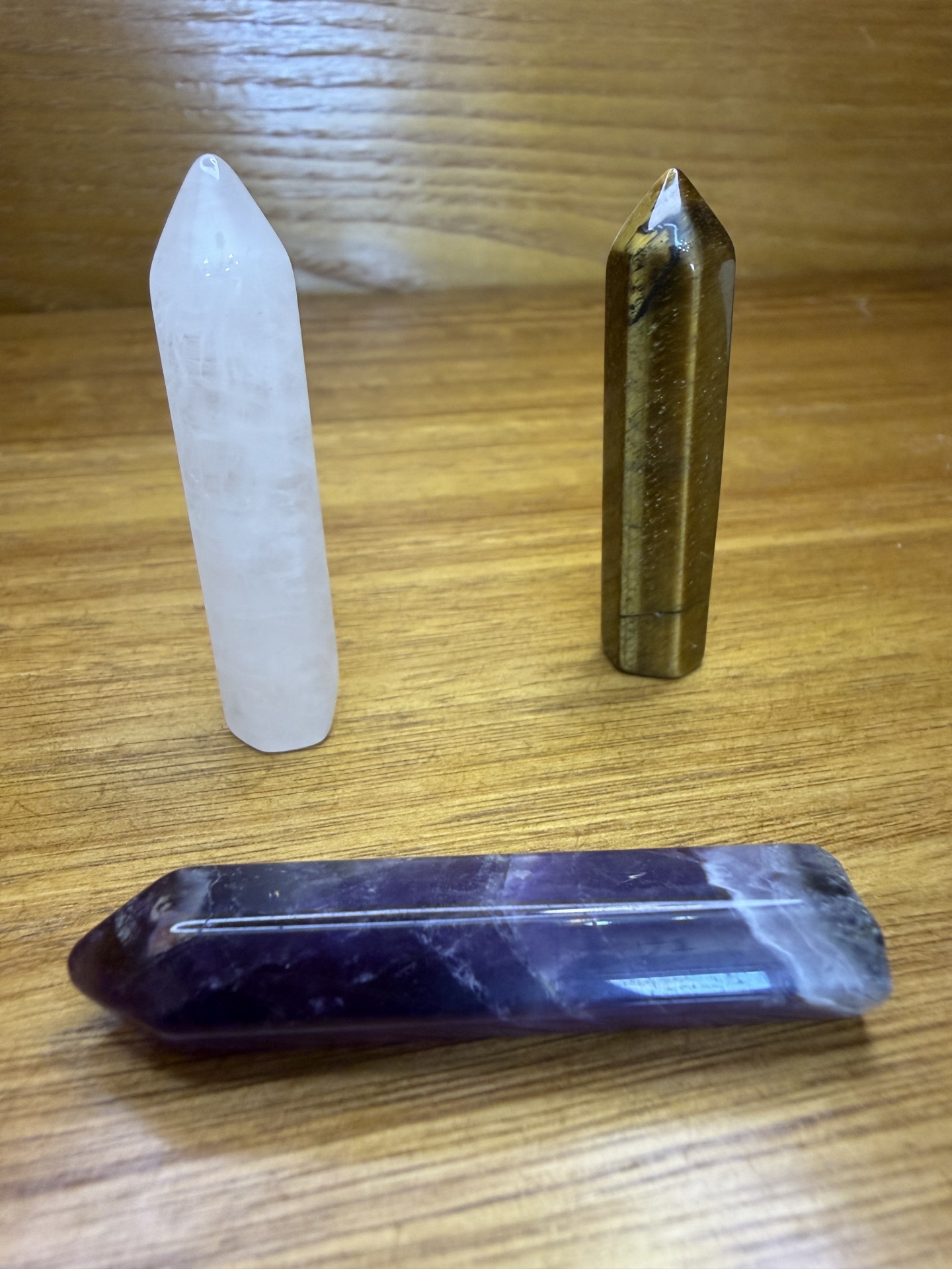 Três lápis de cristal polidos sobre madeira clara, cores branco, castanho e roxo.