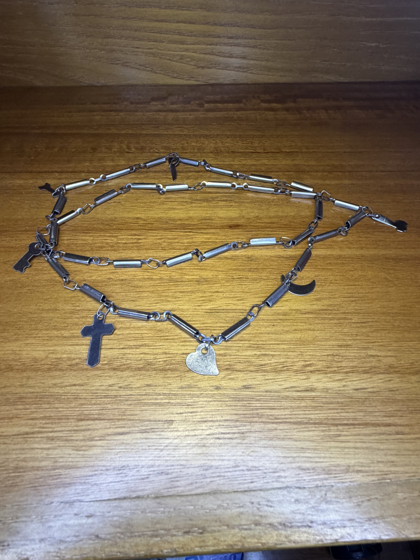Colar de metal prateado com vários pendentes em forma de cruz, coração, chave e meia-lua sobre madeira.