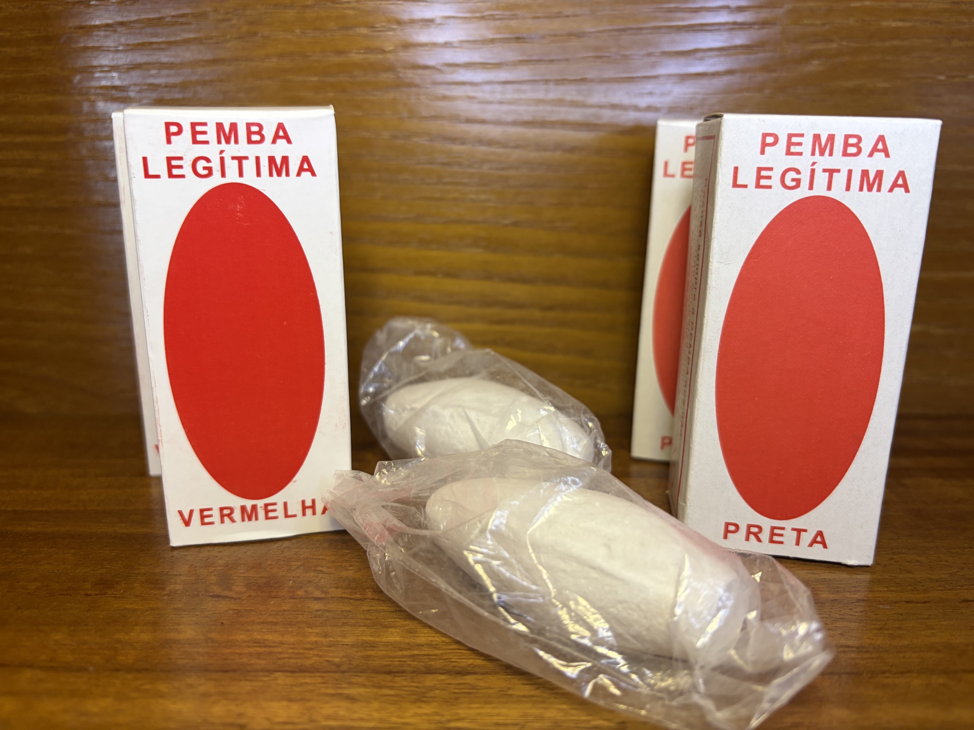 Caixas brancas com oval vermelho e texto PEMBA LEGÍTIMA e objetos brancos embalados em plástico sobre madeira.