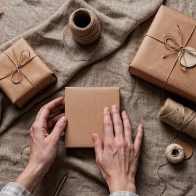 Presentes embalados com papel kraft e corda natural sobre tecido bege, mãos seguram um presente