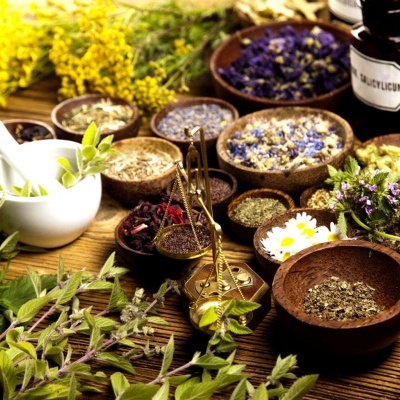 Ervas secas, flores, frascos de vidro, pilão branco e balança dourada sobre mesa de madeira.