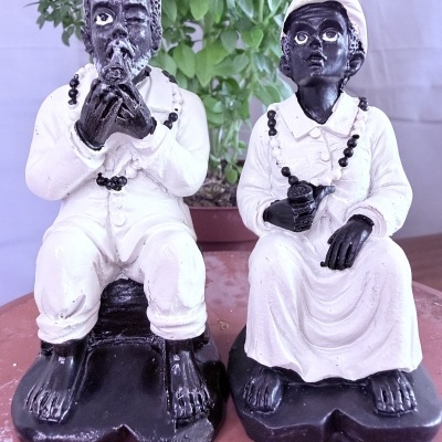Esculturas em preto e branco de duas figuras humanas sentadas com colares, em frente a planta verde.