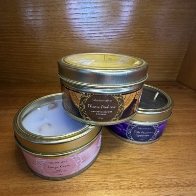 Três velas aromáticas em latas metálicas redondas etiquetadas em português sobre superfície de madeira