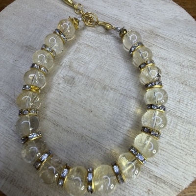 Pulseira de contas transparentes amareladas com aros dourados e pedras brilhantes sobre base de madeira clara