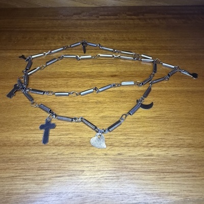 Colar de metal prateado com vários pendentes em forma de cruz, coração, chave e meia-lua sobre madeira.