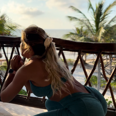 Mulher a fazer agachamento em roupa desportiva azul-escura com presilha branca no cabelo, na varanda perto da praia.