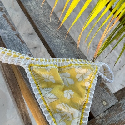 Calcinha de biquíni amarela com estampa de folhas brancas e bordado branco, sobre madeira rústica com folhas verdes.