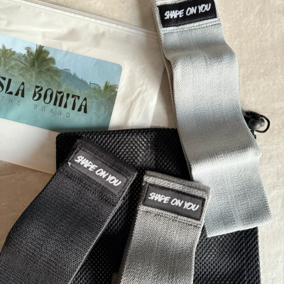 Bandas elásticas para exercício em cinza claro, cinza escuro e preto com etiqueta SHAPE ON YOU, saco de malha preta e embalagem branca com autocolante ISLA BONITA.