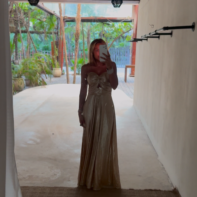 Mulher com vestido dourado comprido a tirar selfie em ambiente com teto de ripas de madeira e plantas