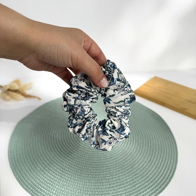 Elástico de cabelo scrunchie branco com padrão floral azul sendo segurado por uma mão.