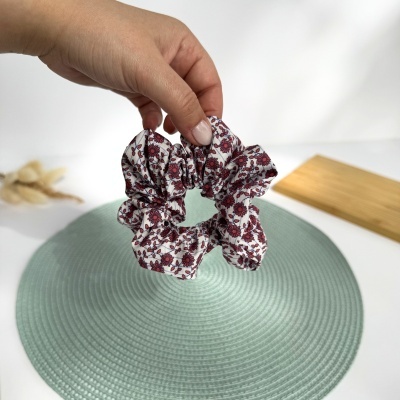 Scrunchie branco com flores vermelhas segurado por mão sobre suporte redondo verde