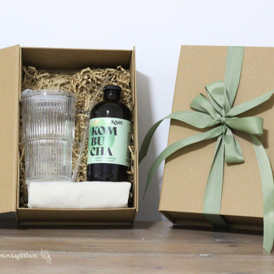 Conjunto de presente com caixa de cartão, copo transparente, garrafa de kombucha e pano branco.