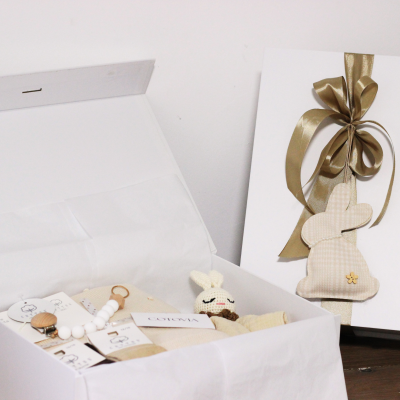 Caixa branca com itens para bebé e decoração com fita dourada e coelho de tecido.