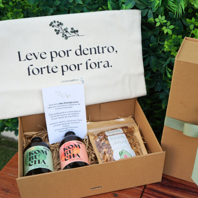Caixa aberta com bebidas kombucha, granola, cartão com texto e toalha com frase motivacional