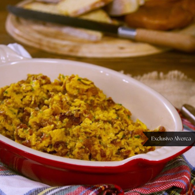 Prato vermelho com mistura amarelada e castanha sobre pano xadrez e tábua com pão ao fundo