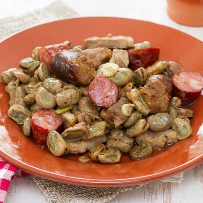 Prato com guisado de feijão fava, chouriço e carne sobre mesa branca com guardanapo às riscas e talheres