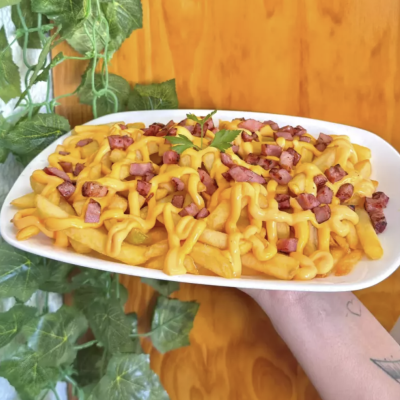 Prato branco com batatas fritas cobertas com molho amarelo e pedaços de bacon, decorado com folhas verdes e fundo de madeira clara.