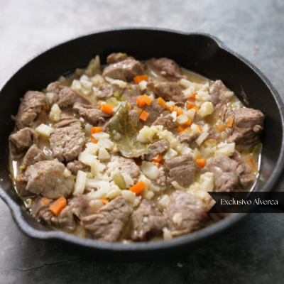 Frigideira preta com carne, cenoura, cebola e folha de louro em molho claro.