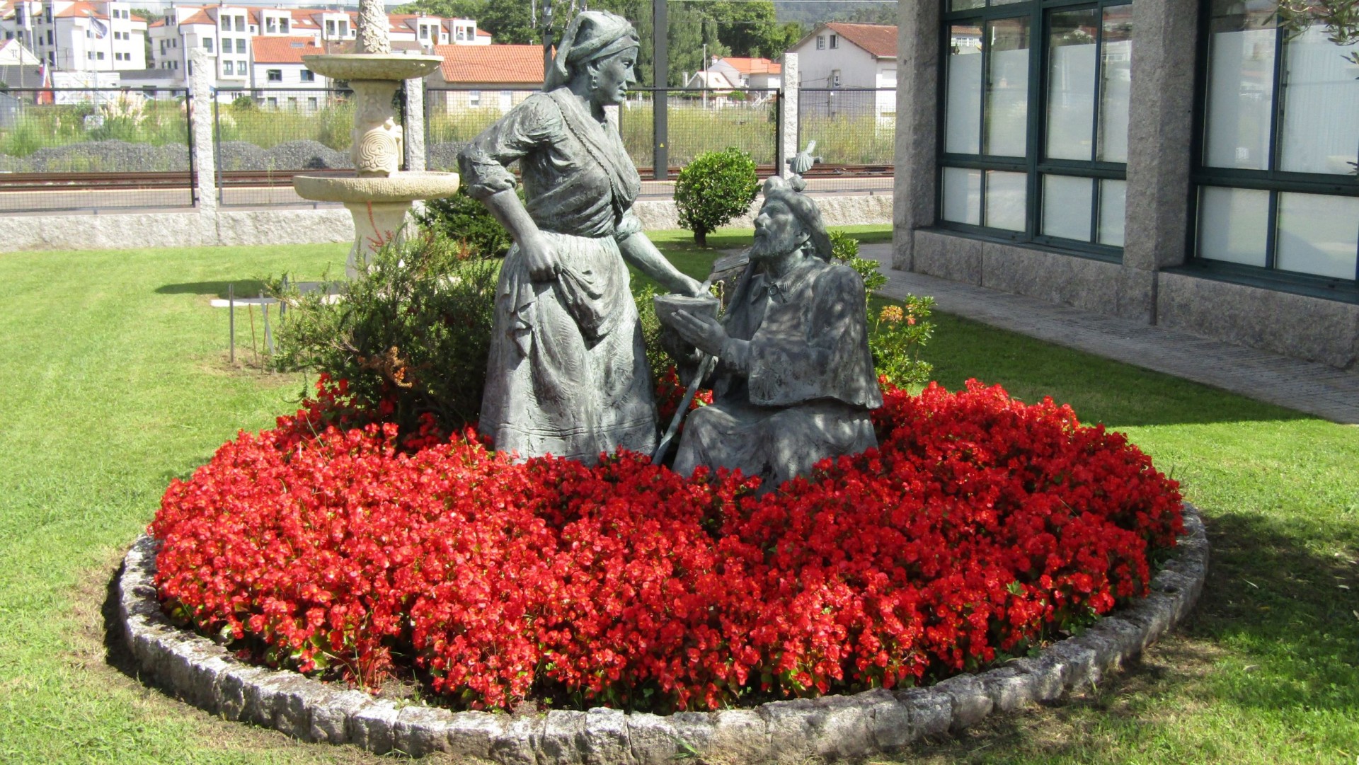 Escultura de duas figuras humanas em metal num canteiro de flores vermelhas num jardim