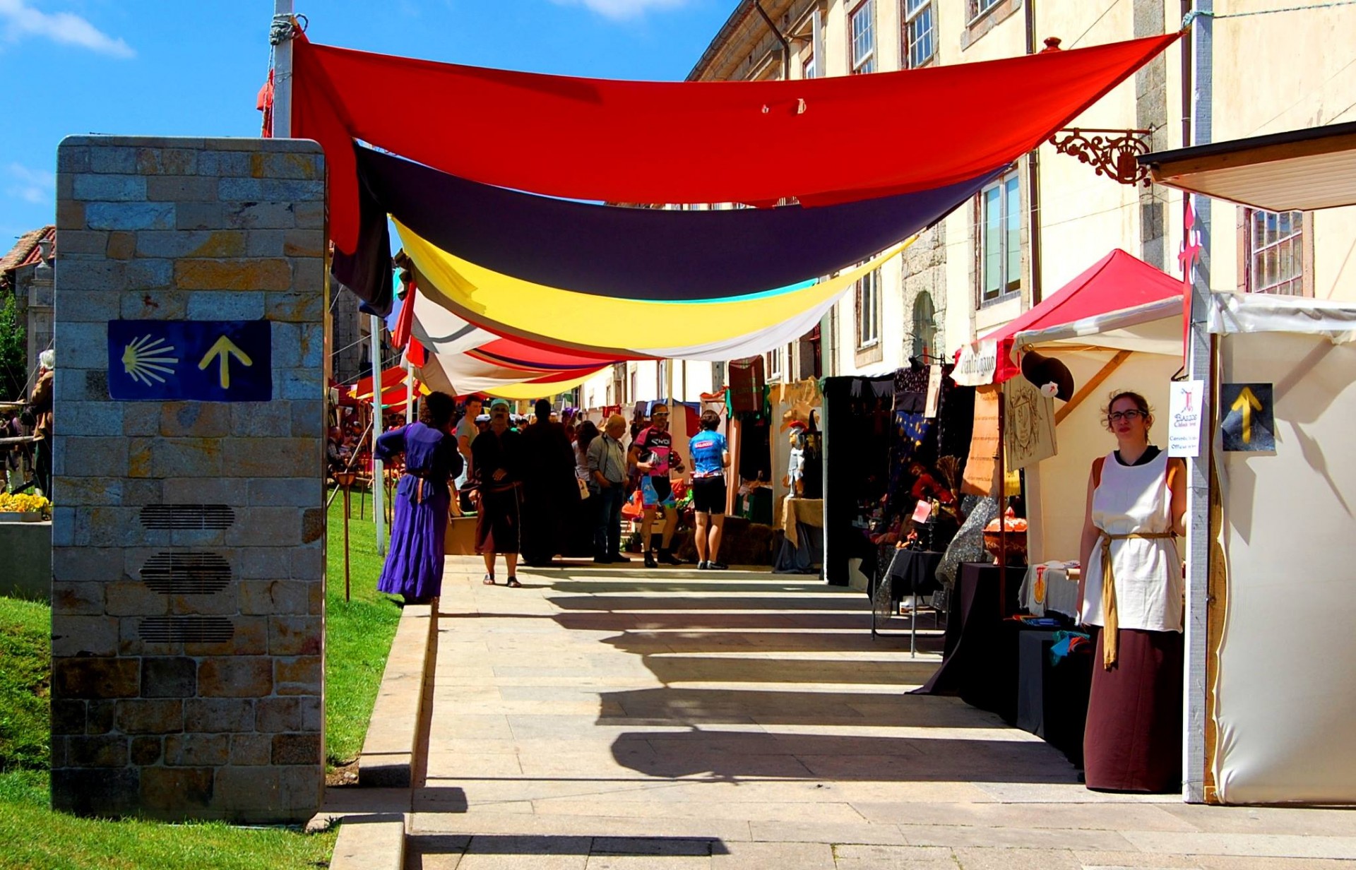Bancas de feira ao ar livre com toldos coloridos e uma placa de direção com concha amarela.
