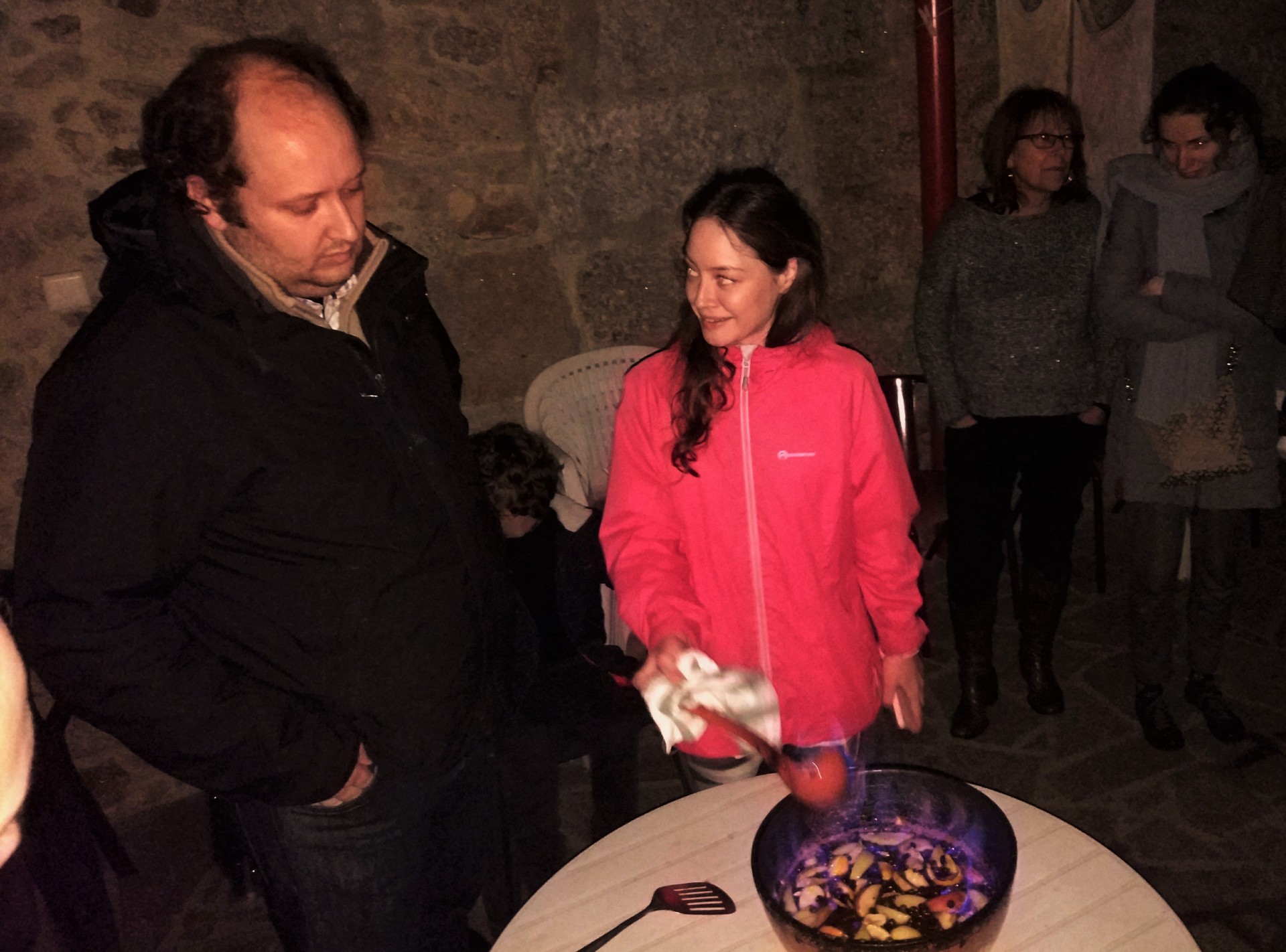 Homem e mulher junto a uma mesa com recipiente azul com comida numa sala com paredes de pedra