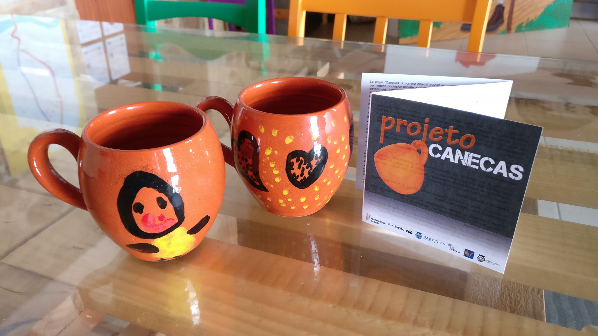 Duas canecas laranja pintadas à mão e cartão com texto 'projeto CANECAS' numa mesa de madeira.