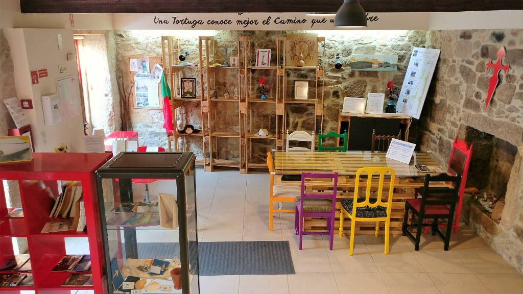 Espaço interior com mesa de madeira e cadeiras coloridas, estantes de madeira com objetos, paredes de pedra