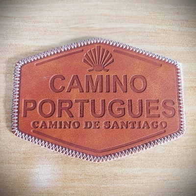 Placa de couro castanho com texto gravado 'CAMINO PORTUGUES CAMINO DE SANTIAGO' e bordas cosidas brancas