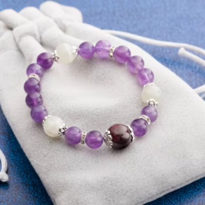 Pulseira elástica de contas roxas, brancas e granada com espaçadores prateados em saco de veludo branco.