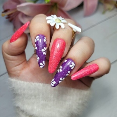 Unhas postiças rosa e roxas com flores brancas e bolinhas, sobre fundo branco com flores naturais.