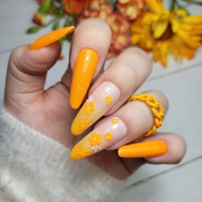 Unhas compridas com esmalte laranja e flores decorativas, anel amarelo e fundo com flores.